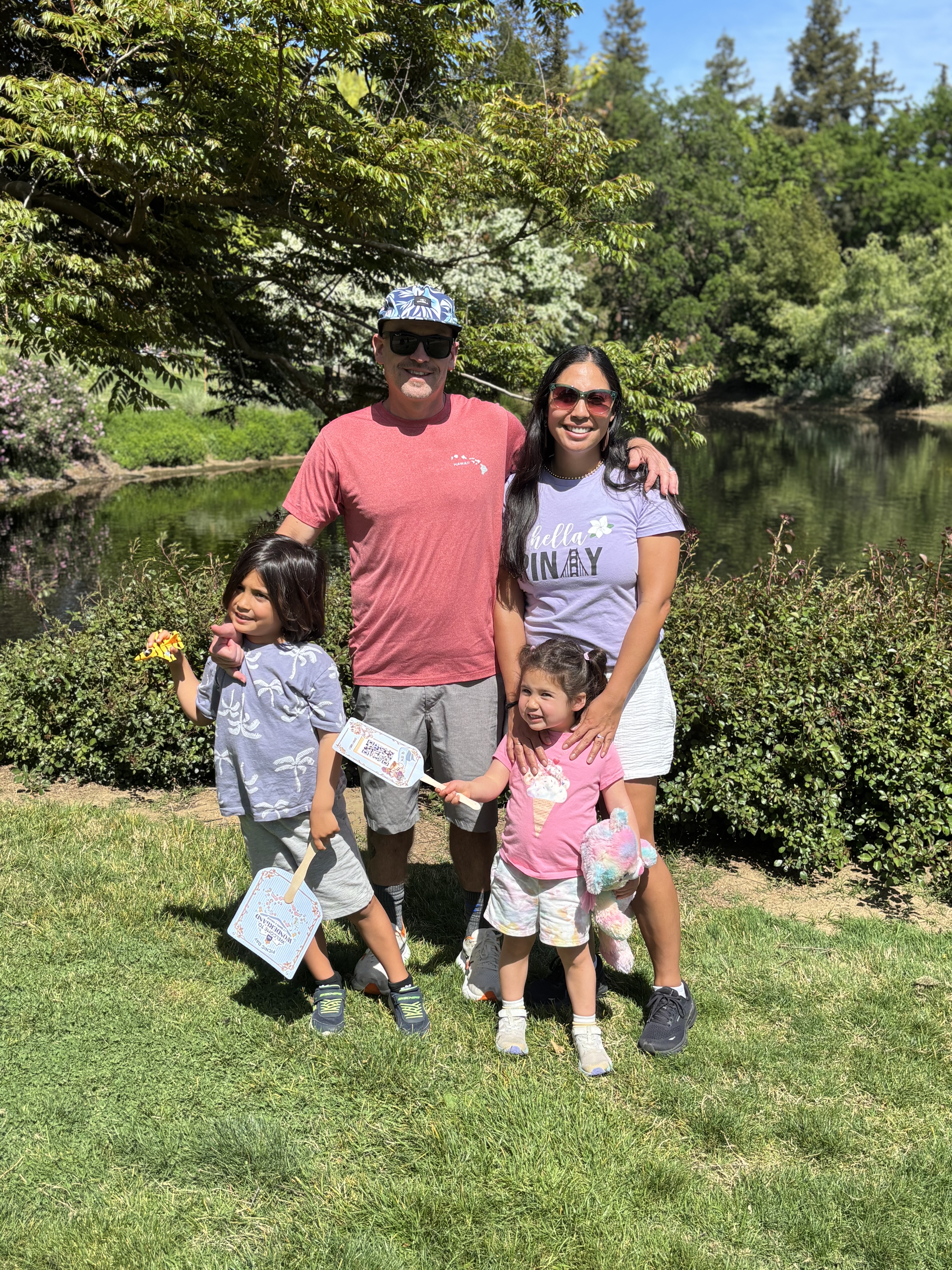 Family of four—parents and two young daughters—smiling by a grassy lakeside