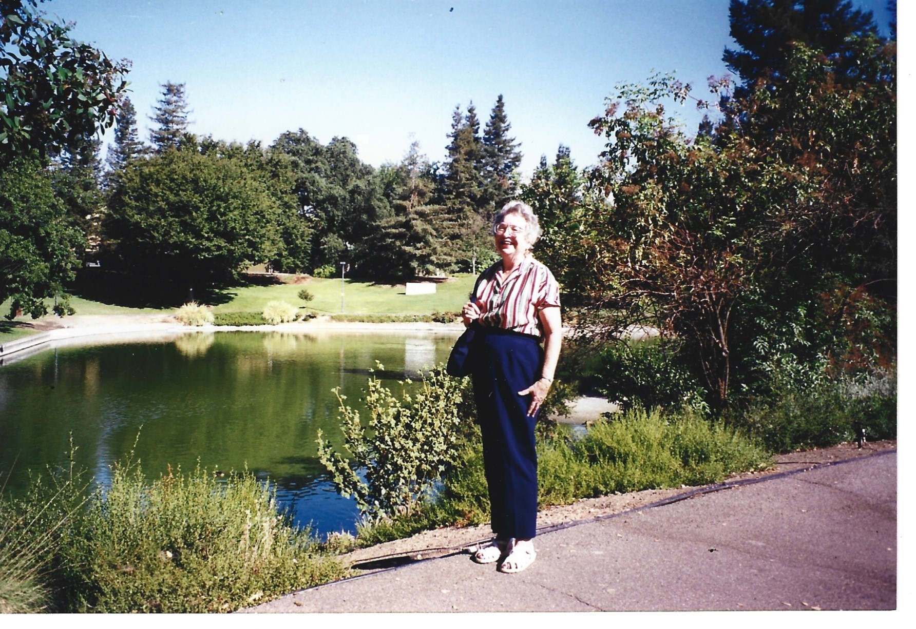 Woman standing on a sunlit park path beside a calm pond, trees and shrubs.