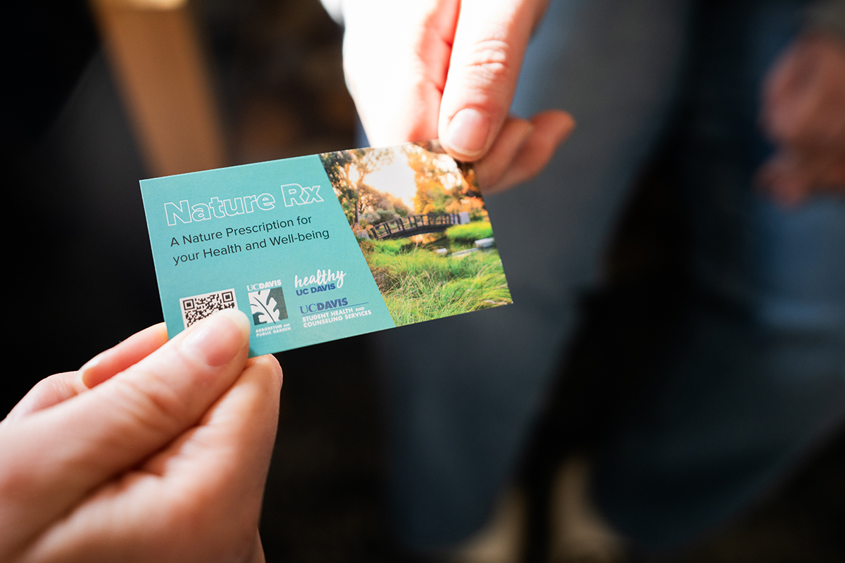 Image of a hand giving another hand a business-type card with a beautiful outdoor image of a bridge in the UC Davis Arboretum and includes the copy:  Nature Rx A Nature Prescription for your Health and Well-being. The card includes a QR code and wordmarks from the participating partners including the UC Davis Arboretum and Public Garden, Healthy UC Davis, and UC Davis Health and Counseling Services.