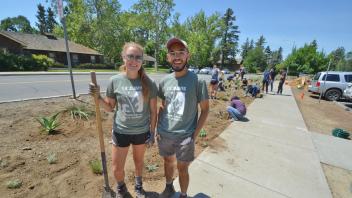 In the foreground, two student interns smile and pose with a shovel, while in the background, several other students work on planting a landscape on a long patch of dirt between La Rue Road and the sidewalk