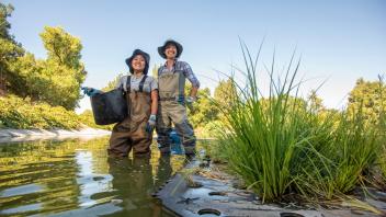 Two students smile, wearing waders and standing in the Arboretum Waterway