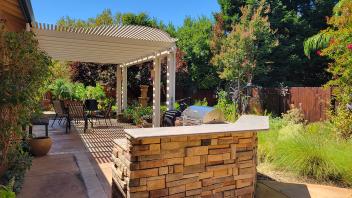 Mary Kay Williams backyard garden showing a paved and shaded patio area with a grill and chairs surrounded by lush green plants and bright red flowers.