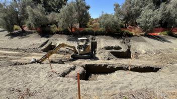 The drained waterway shows an expanse of dirt, an excavator is in the middle digging holes throughout.