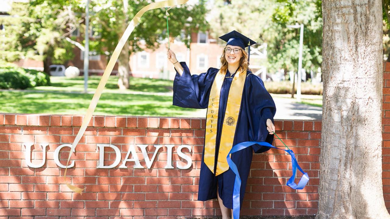 Image of UC Davis grad in cap, gown and stole waving blue and gold streamers in front of the brick UC Davis monument.