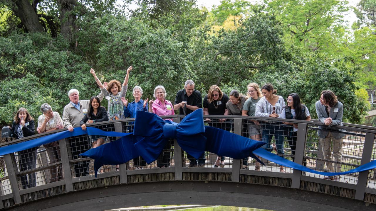 Image of a line of people on an arched bridge celebrating by cutting a large blue ribbon on the side of the bridge railing.