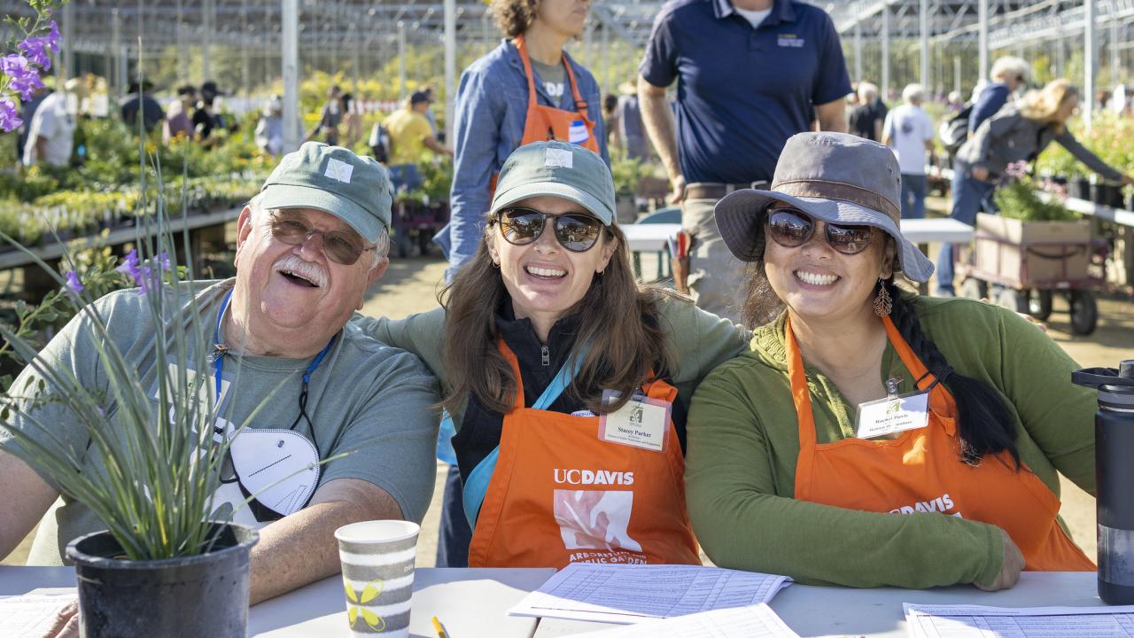 An Arboretum volunteer and two staff members smile, sitting at a table at an Arboretum Plant Sale