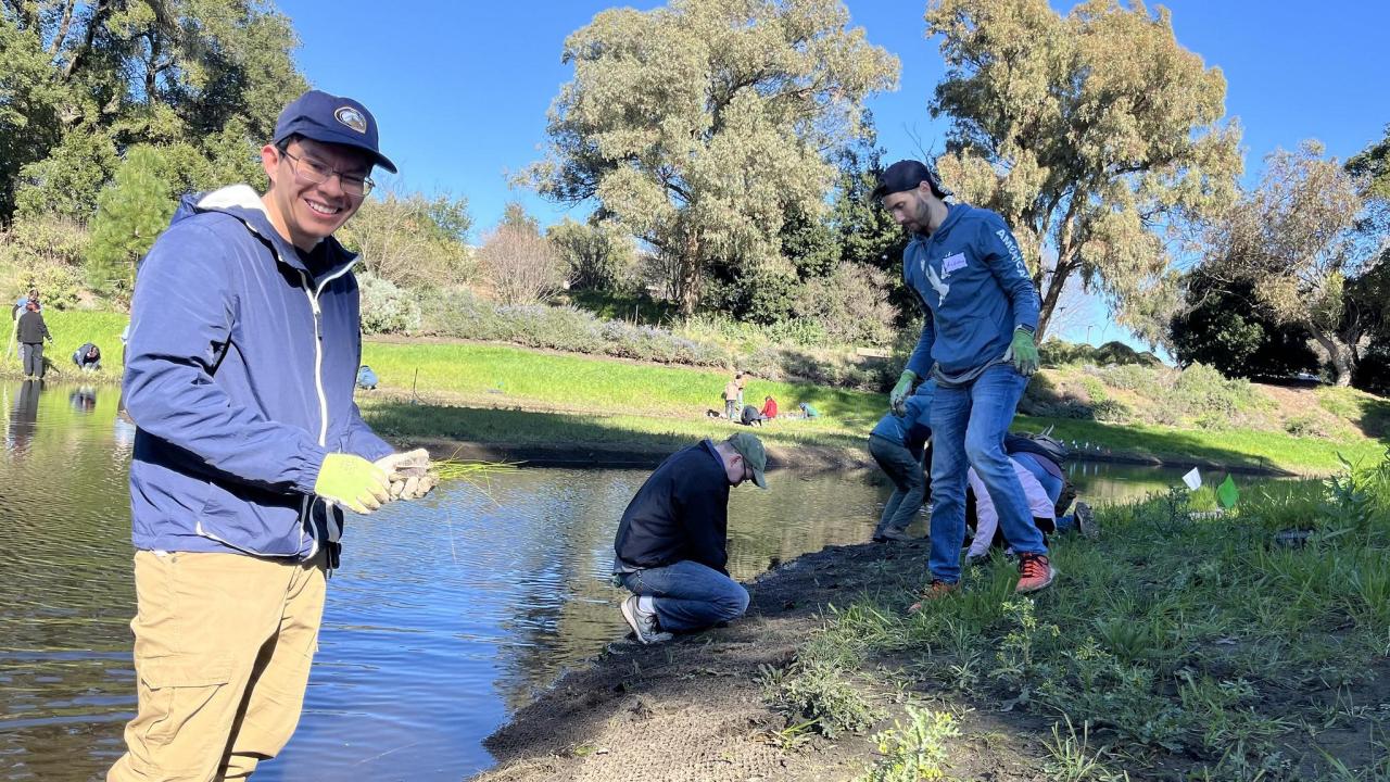 One Arboretum student intern smiles in the foreground while a dozen volunteers work on the banks of the Arboretum Waterway in the background