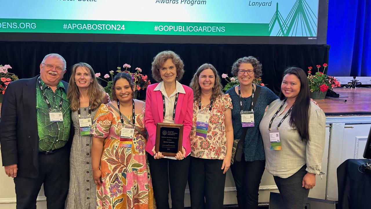 Kathleen Socolofsky holds an award, standing in the center of a group of seven people in front of a stage.
