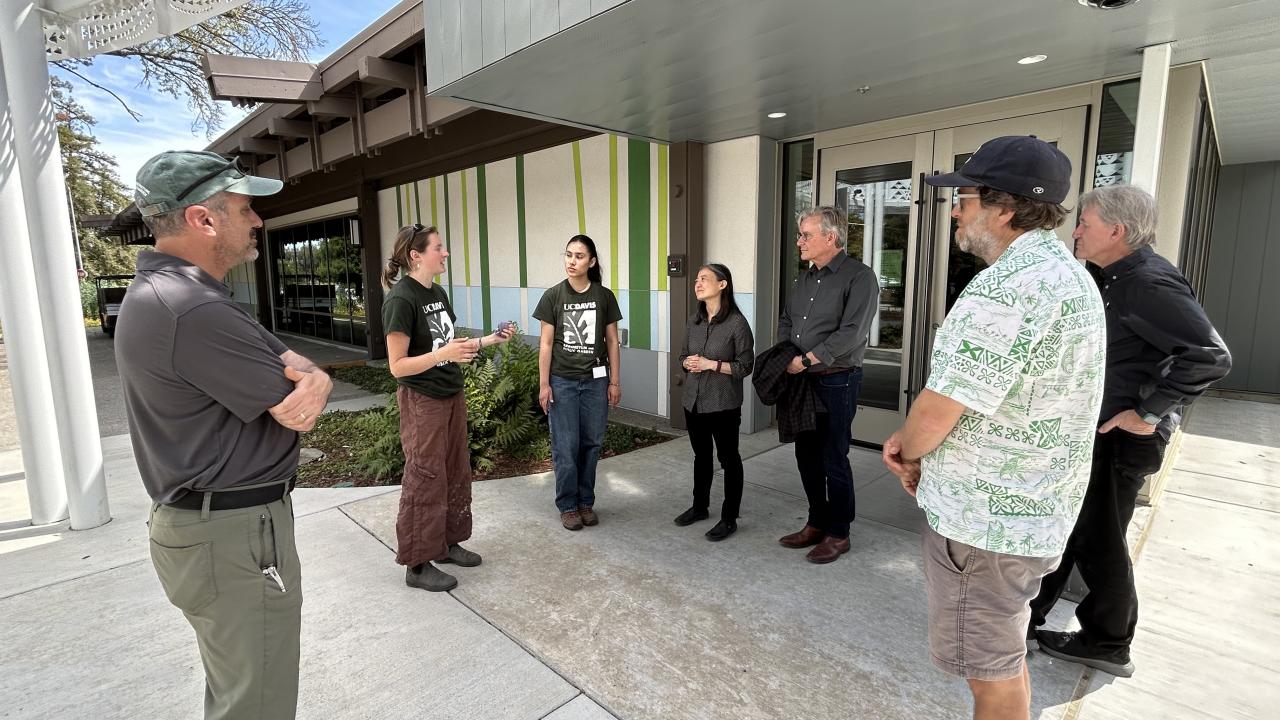 Group of six accreditation team members standing outdoors near a building, with two students, Dyanna Perez and Amelia Swanson, explaining something to the group in front of a modern, green-accented building.