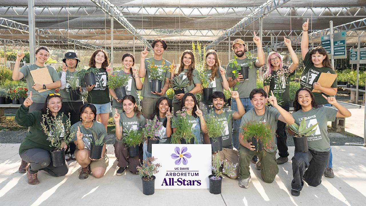 A group of UC Davis Arboretum and Public Garden students and staff pose together at the Teaching Nursery, smiling and holding potted plants. They wear matching green shirts and raise their fingers to signal “number one” in celebration of the Arboretum All-Stars program. A sign with the UC Davis Arboretum All-Stars logo is displayed in front of the group. Rows of plants and nursery shade structures are visible in the background.