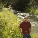 A man in a rust-colored short-sleeve shirt stands smiling beside a lush, sunlit stream surrounded by tall green grasses and foliage. Dappled sunlight sparkles on the water behind him, and dense trees line the shaded background.