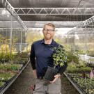 Image of UC Davis Arboretum and Public Garden Nursery Manager Taylor Lewis standing among the aisles of the UC Davis Arboretum Teaching Nursery, smiling and holding a flowering maple in one hand with a hand positioned on the opposite hip. Taylor has red hair, a beard, wears glasses and dons a welcoming smile. He is missed terribly.