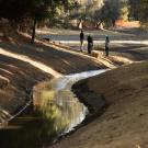 In the foreground, a narrow, curving portion of the Arboretum Waterway is shown with water while three people look in the opposite direction to a larger area of the Waterway.