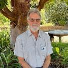 man with glasses wearing collared blue and green striped shirt sitting with tree and green foliage in background. a circular table sits in the background
