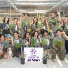 A group of UC Davis Arboretum and Public Garden students and staff pose together at the Teaching Nursery, smiling and holding potted plants. They wear matching green shirts and raise their fingers to signal “number one” in celebration of the Arboretum All-Stars program. A sign with the UC Davis Arboretum All-Stars logo is displayed in front of the group. Rows of plants and nursery shade structures are visible in the background.