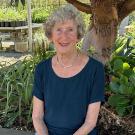 woman sitting down wearing navy blue blouse. green foliage and tree behind her