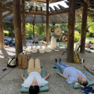 People lying on mats under wooden pergola in outdoor relaxation session with singing bowls